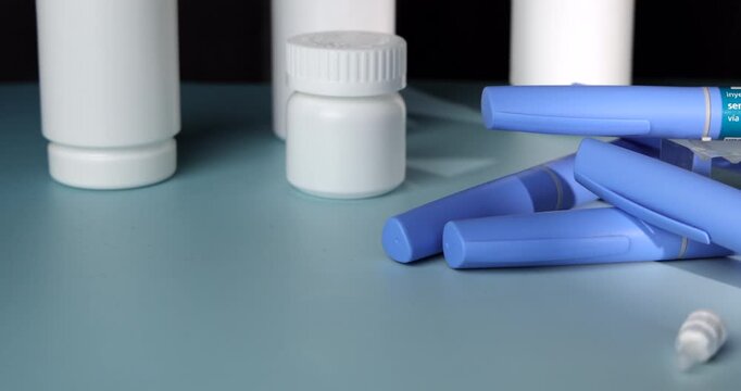 Horizontal tracking camera movement passing along blue Semaglutide GLP-1 injection pens placed near white pill bottles on a light blue table surface over a dark black background, healthcare setup