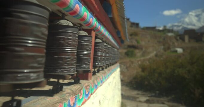 Annapurna Circuit, Manang, Nepal, gliding shot along a row of traditional metal Buddhist prayer wheels installed on a painted stone wall in Manang village.