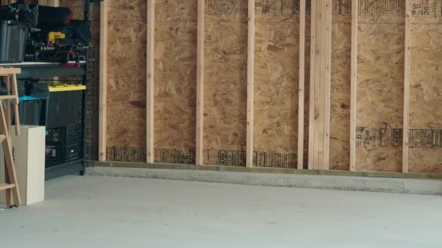 Static shot of an unfinished garage wall with exposed plywood, shelving full of camera equipment, and a concrete floor