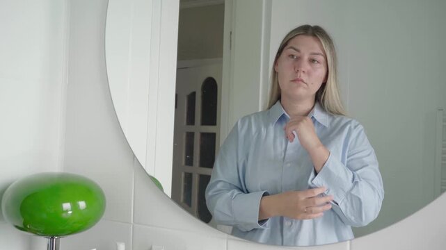 Young woman in a bathroom adjusts her shirt cuffs while looking at herself in the mirror before an online job interview.