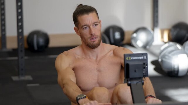 Medium close-up of a shirtless athletic man with a man bun turning his head while holding a Concept2 rower handle in a busy gym warmup area.
