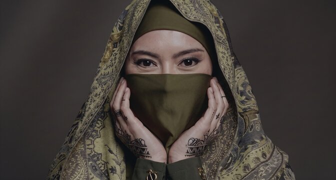Close up portrait of a young Muslim woman wearing a traditional headscarf and niqab covering her face against a dark background