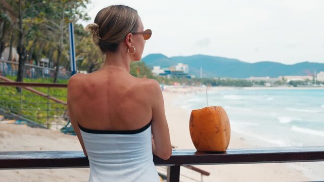 Blonde woman enjoying beautiful sea view and coconut cocktail from beach bar. Rich female vacationer admiring ocean panorama from coastline, back view, relaxed atmosphere in summer holidays, traveling