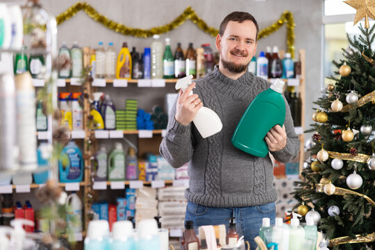 Bearded man in knitted sweater smiling while comparing two cleaning product bottles in home care store decorated for Christmas, making thoughtful purchase decision