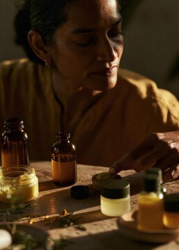 Young woman sitting at a wooden table with several small bottles and jars of essential oils scattered around her. she is wearing a yellow shirt and has her hair pulled back in a bun.