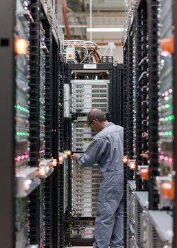 Man in a server room, working on a rack of servers. he is wearing a blue coverall and is standing in front of a large server rack with multiple rows of servers and cables.
