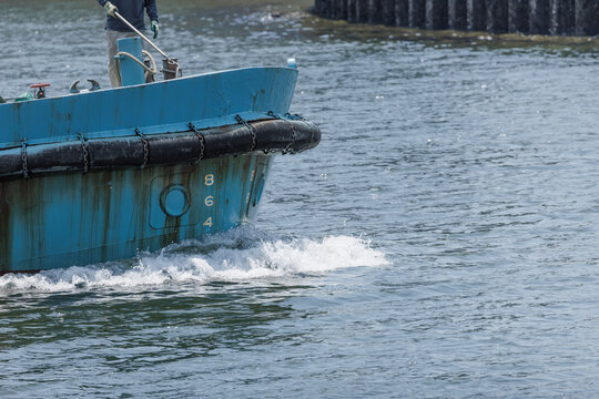 Blue industrial workboat with cylindrical rubber fenders and draft marks moving through harbor water as a crew member handles a boat hook.