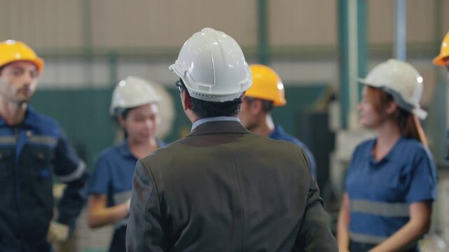 Man in white hard hat addresses group. Workers wear safety gear, listen attentively. Team appears engaged, focused on instruction