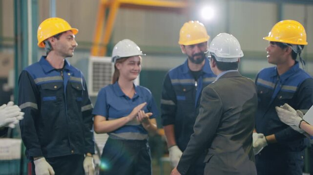 Factory workers wear hard hats and gloves. They exchange smiles and gestures with a visiting manager. Team members show enthusiasm