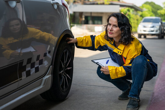 female auto mechanic holding clipboard,sitting and checking car tire at outdoors repair shop garage