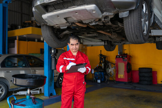 expertise asian senior mechanic in red uniform writing on clipboard while inspecting a car on hydraulic lift in auto repair shop