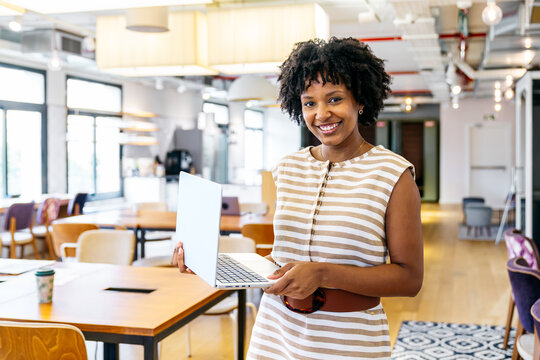 Young black businesswoman smiling, holding a laptop, working remotely in a contemporary shared office space with natural light