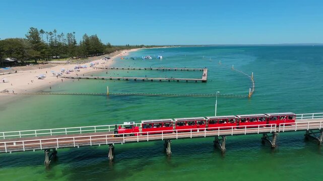 Aerial view of the iconic Busselton Jetty with its red train traveling over the turquoise waters of Geographe Bay near the sandy beach in Busselton, Western Australia, Australia.