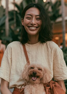 Portrait of a young woman with shoulder-length dark hair, smiling widely and laughing. she is wearing a beige t-shirt and brown pants, and is carrying a small pink dog in her arms.