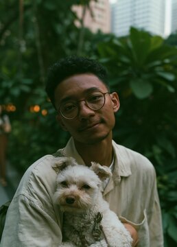 Portrait of a young man holding a small white dog in his arms. the man is wearing a beige shirt and glasses, and he is smiling at the camera.