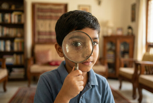 Curious Indian boy holding magnifying glass to eye for exploration in warm natural light home library interior