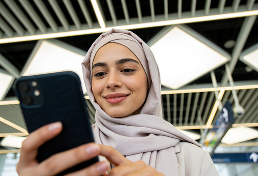 Smiling young Muslim woman wearing beige hijab using smartphone in modern airport terminal under bright lights