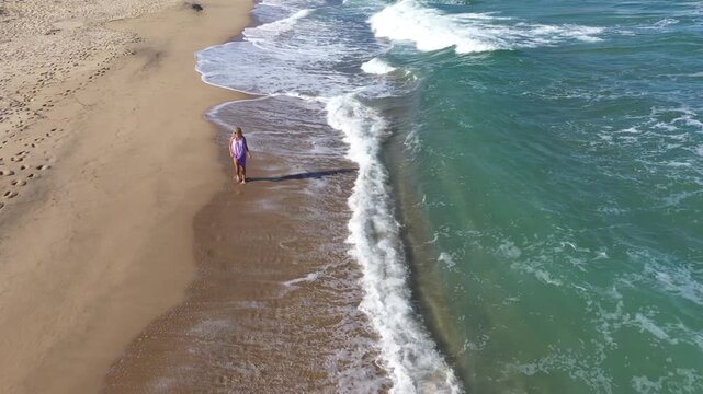 Slow motion aerial view of a gorgeous blonde woman walking on a beach. Sea waves washing sandy shoreline