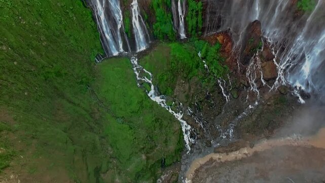 Aerial view of Tumpak Sewu Waterfall with cascading water, lush green cliffs, and rocky riverbed in Kecamatan Pronojiwo, Jawa Timur, Indonesia.
