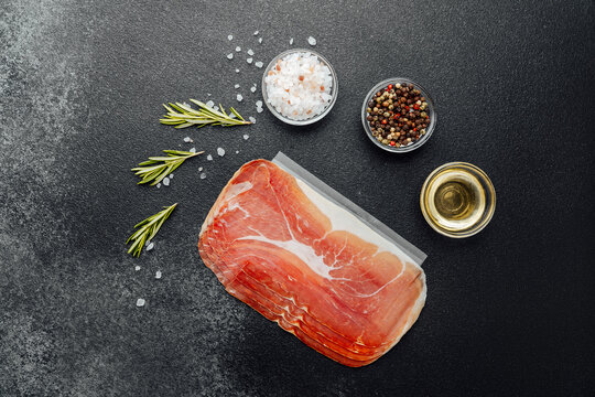 Pack of sliced prosciutto ham with salt, pepper and herbs on black background