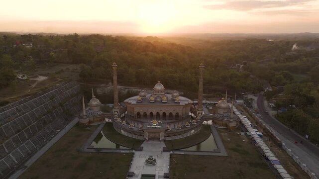 Aerial view of the Masjid Samin Baitul Muttaqin featuring golden domes and minarets surrounded by lush greenery during sunset in Bojonegoro, East Java, Indonesia.