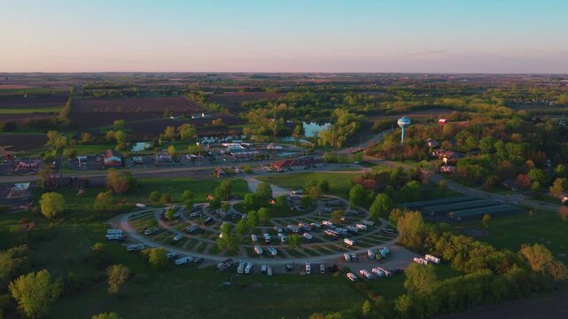 Golden hour drone view of a rural campground near DeForest, Wisconsin, rows of RVs on a circular loop surrounded by farmland, trees and a small pond at sunset.