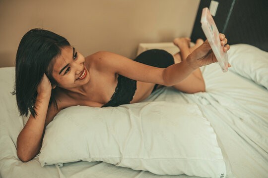 Smiling adult woman in lingerie takes a playful smartphone selfie from bed, suggesting romantic sharing, social media posting, confidence, and digital lifestyle.