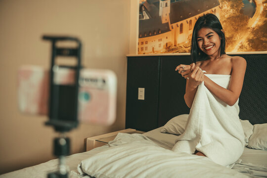 Smiling adult woman wrapped in a towel poses for a smartphone camera in bed, creating premium video content for online subscribers and fans.