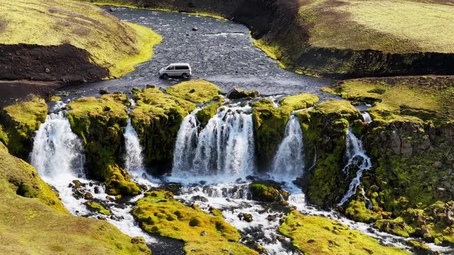 Aerial view of Blafjallafoss waterfall with a van crossing the river through the mossy green landscape of Kirkjubaejarklaustur, Skaftarhreppur, Iceland.