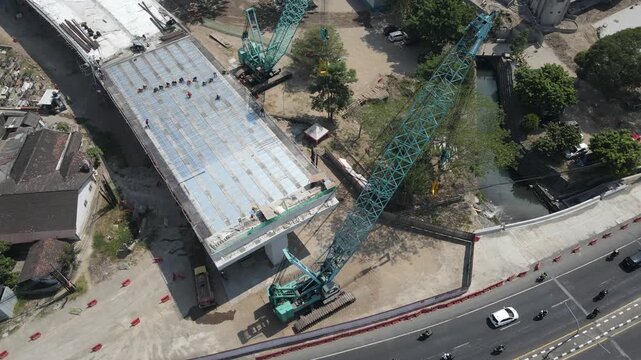 Aerial view of a large crawler crane working on a concrete bridge deck for flyover girder construction site with workers and nearby road traffic in Special Region of Yogyakarta, Indonesia.