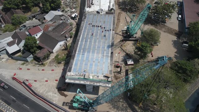 Aerial view of a large crawler crane working on a concrete bridge deck for flyover girder construction site with workers and nearby road traffic in Special Region of Yogyakarta, Indonesia.