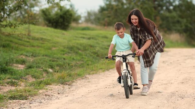 Warm mother and son bicycle lesson unfolds on sandy village road under cloudy skies. Boy squints when riding becomes difficult yet both share joyful family moment.