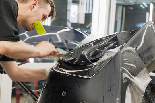 Professional car detailing master wrapping a luxury vehicle front end with clear polyurethane anti-gravel film for paint protection. Close-up of protective PPF wrap application installation service. 