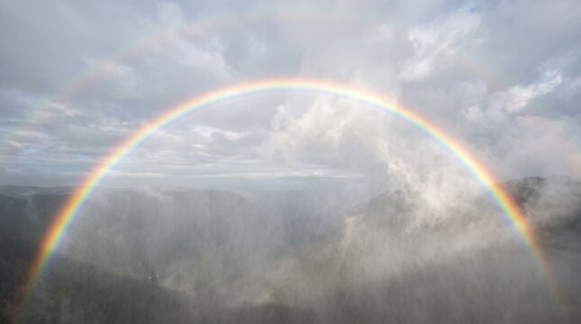 Vibrant full rainbow arch appearing in cloudy sky over misty mountains and forest, atmospheric phenomenon with bright colors, dramatic clouds and fog in valley below, photorealistic, ultra-detailed, 8