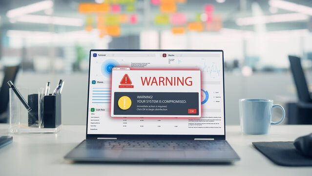 Laptop on Desk Displaying a Critical Virus Alert Warning Pop-up Over Financial Charts, Signaling a Compromised System in Open Plan Office Interior.