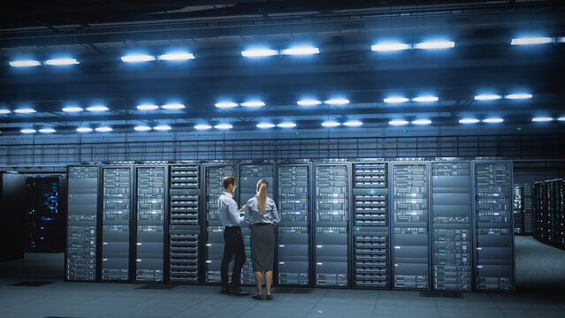 Two Caucasian IT Engineers, Stand With Their Backs Turned Inspecting Server Rack Cabinets in a Vast Modern Data Center Illuminated by Rows of Bright Ceiling Lights