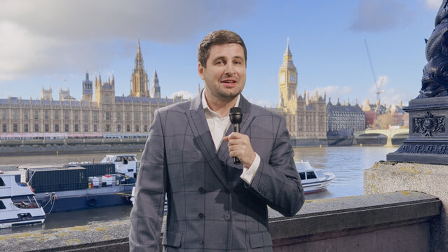 News Reporter Holds a Microphone and Speaks to Camera While Filing an On-location Television Report From the Thames Embankment, With Westminster Palace, Big Ben and Moored Boats Behind