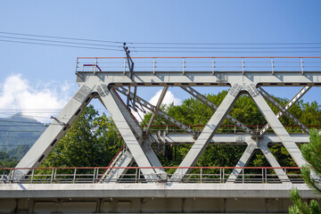 Fototapeta premium Steel truss railway bridge structure over river against blue sky and mountain trees background. Real Photo