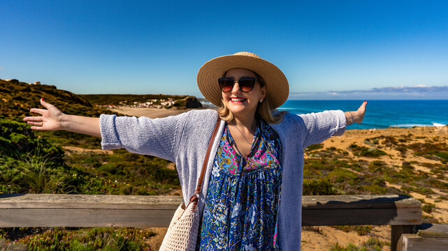 Happy mature woman tourist standing at viewpoint to Monte Clerigo beach on Algarve coast on sunny spring day. Front view
