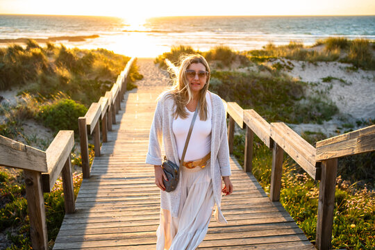 Beautiful middle-aged woman tourist walking up stairs overlooking sandy beach on sunset in spring. Front view. Sightseeing Algarve coast in Portugal
