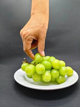 A person's hand picking a fresh green grape from a cluster on a white plate with a black background 