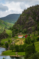 Fototapeta premium Vertical shot of picturesque traditional Norwegian cabins nestled on grassy terraces under a towering rocky cliff by the waters of Lovrafjorden in Suldal, Norway