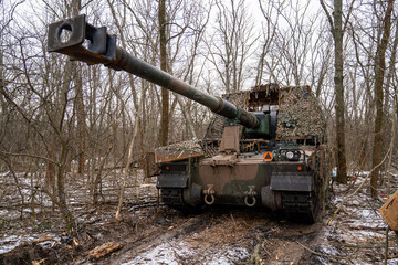 Obraz premium AHS Krab self-propelled howitzer of the Ukrainian Armed Forces camouflaged with a camouflage net and positioned in a winter forest. 
