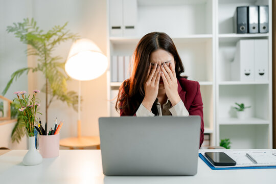 Young asian businesswoman sitting at her desk. Covering her face with hands. Expressing severe stress. Headache. Or burnout while working on a laptop in a modern office