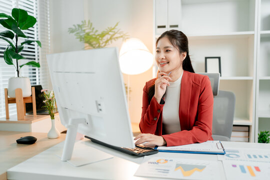 Young asian woman working at a bright modern desk. Smiling confidently while using a computer and keyboard. Surrounded by financial charts and office plants
