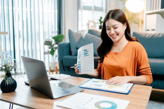 Young asian woman smiling while working from home. Reviewing financial data and analyzing business performance reports with graphs and charts