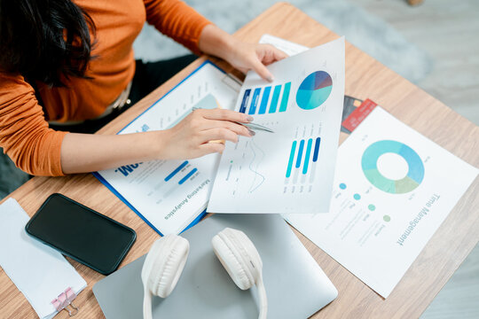 Woman's hands holding a financial report, pointing at charts and graphs while analyzing data and planning business strategy on a desk with a laptop, smartphone, and headphones
