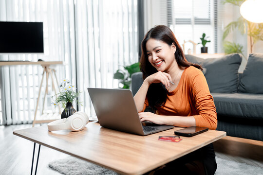 Young asian woman happily engaging with laptop, smiling while typing, using a credit card for convenient online shopping or banking from her comfortable home