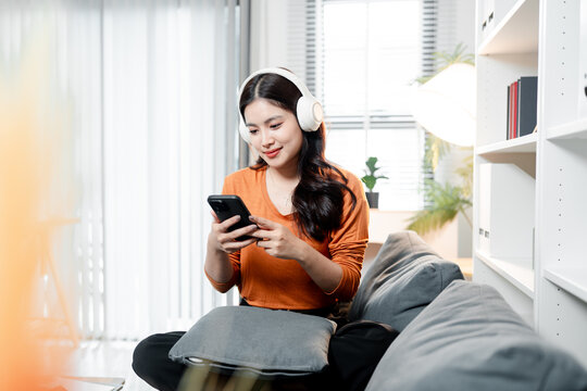Young asian woman wearing white wireless headphones and smiling, listening to music or podcast, enjoying leisure time and digital entertainment at home using smartphone