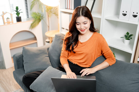 Young asian woman enjoying comfortable remote work at home, sitting on sofa, typing on laptop, smiling while focusing on online tasks, distance learning, or browsing internet